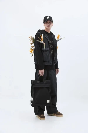 Young man in dark streetwear holding a black tote bag against a white seamless background for Emporio Armani