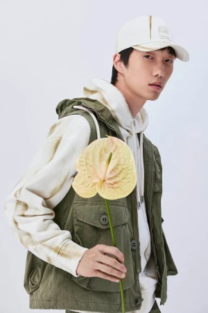 East Asian male model wearing an olive green utility vest and white baseball cap, holding a pale yellow anthurium flower against white.