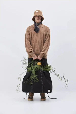 Model wearing a brown bucket hat and patterned sweater holding a large black bag filled with foliage against white