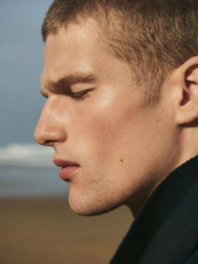 Close-up profile portrait of a male model with short hair wearing a dark high-neck garment against an outdoor coastal background.