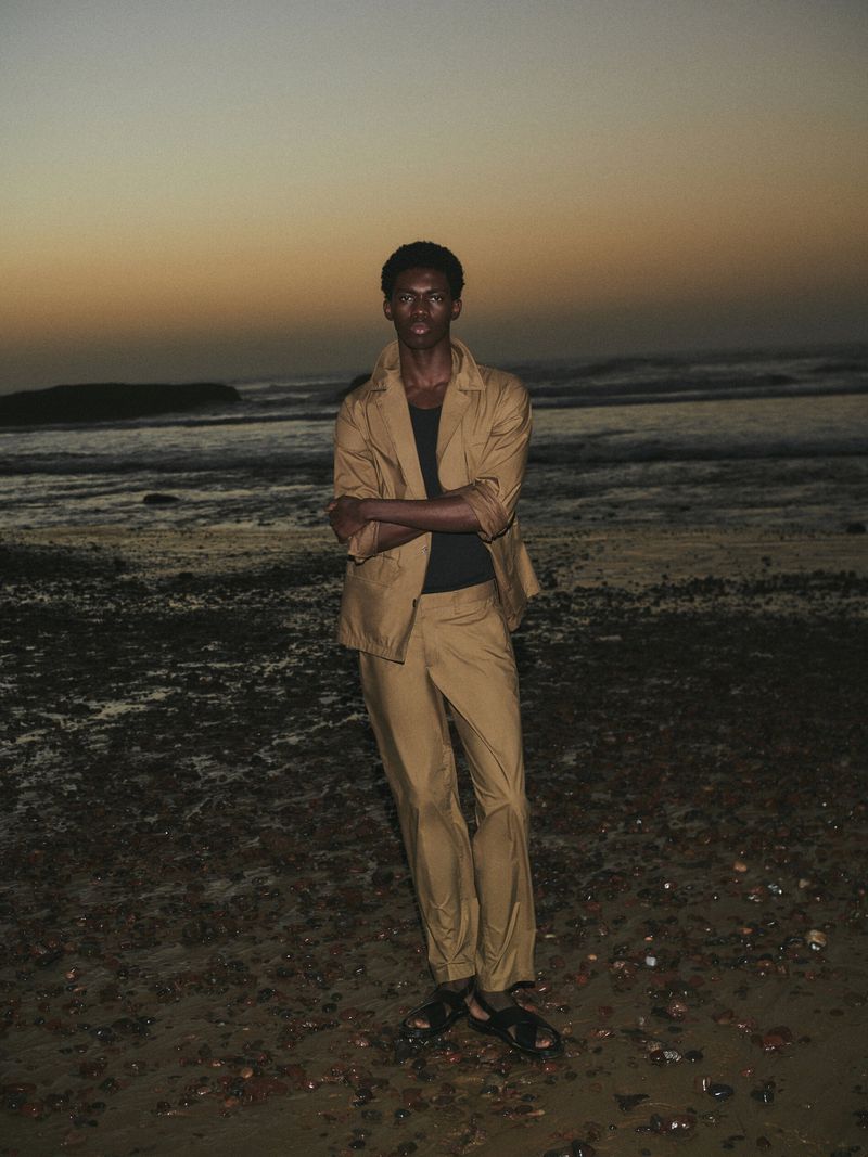 A young Black male model in a tan suit poses on a sandy beach at dusk with the ocean and a darkening sky visible in the background.