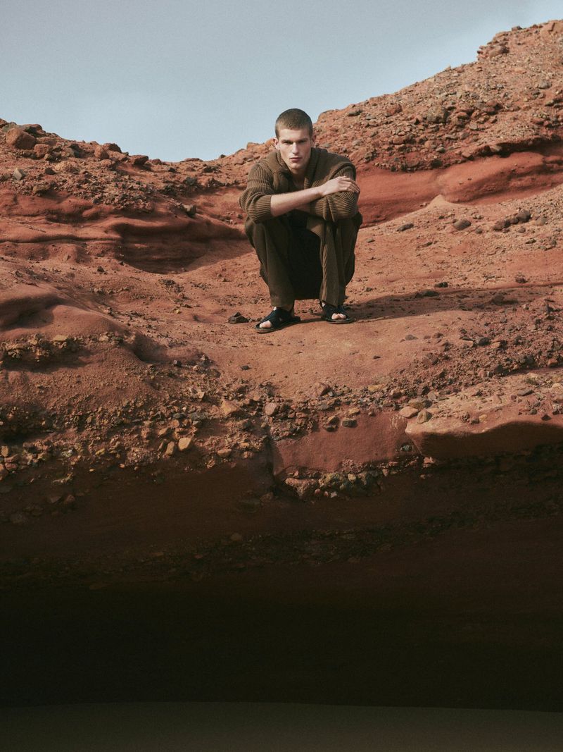 A young man in brown clothing poses in a crouched position in a barren, rocky landscape with a small body of water at the bottom.