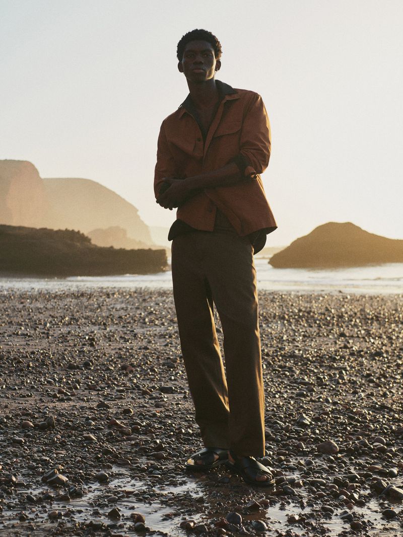 Young man in a copper shirt and brown pants standing on a rocky beach at sunset with large rock formations in the background.