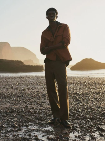Young man in a copper shirt and brown pants standing on a rocky beach at sunset with large rock formations in the background.