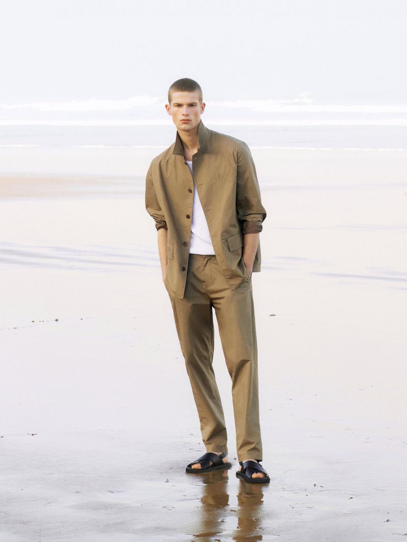 A young man stands on a beach wearing a tan suit jacket and pants. The neutral color palette complements the natural beach environment.
