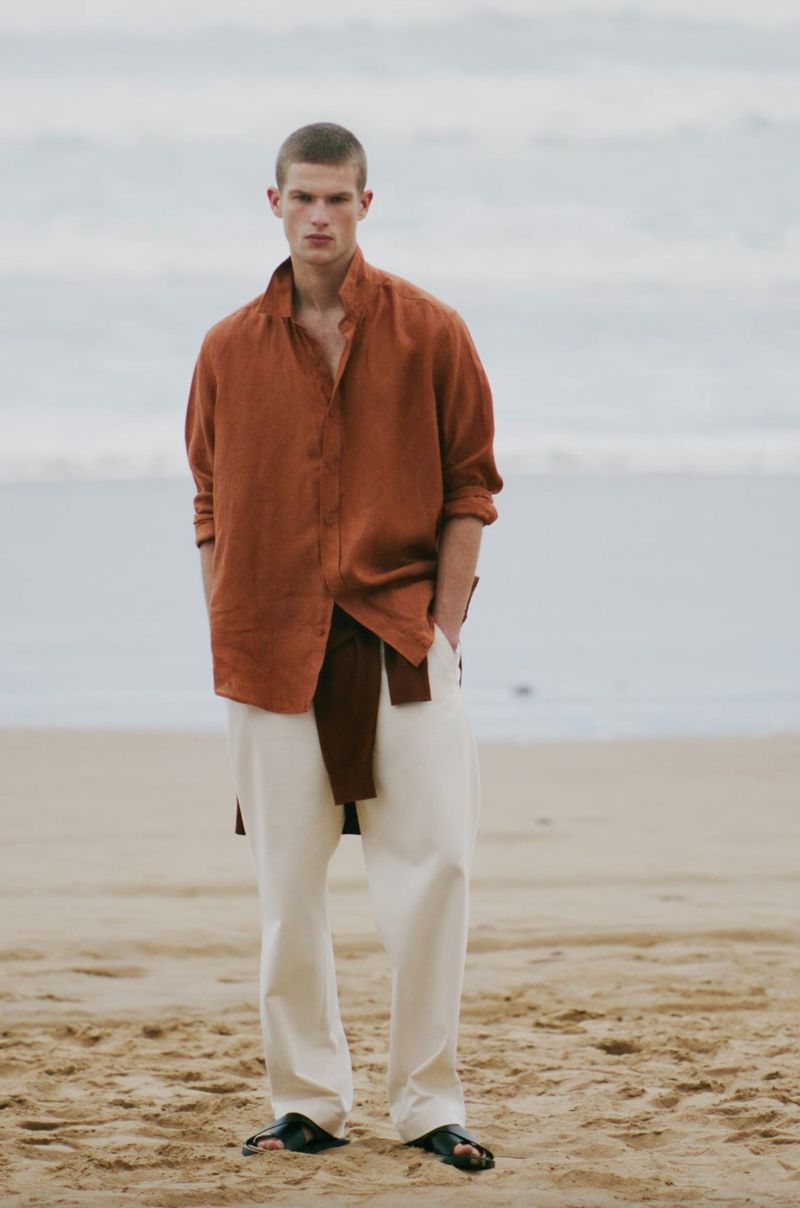 A male model poses on a beach, wearing a burnt orange button-down shirt, cream-colored trousers, and black sandals, against a neutral background.