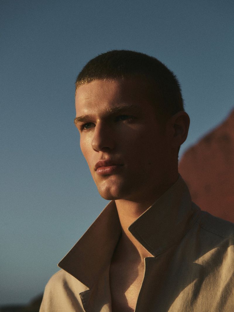 Headshot of a young adult male with short hair wearing a light colored open collared shirt against a blue sky and neutral wall background.