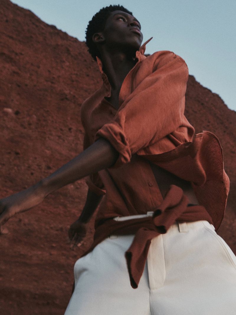 A Black male fashion model poses in an orange button-down shirt and white pants against a reddish-brown hillside backdrop.