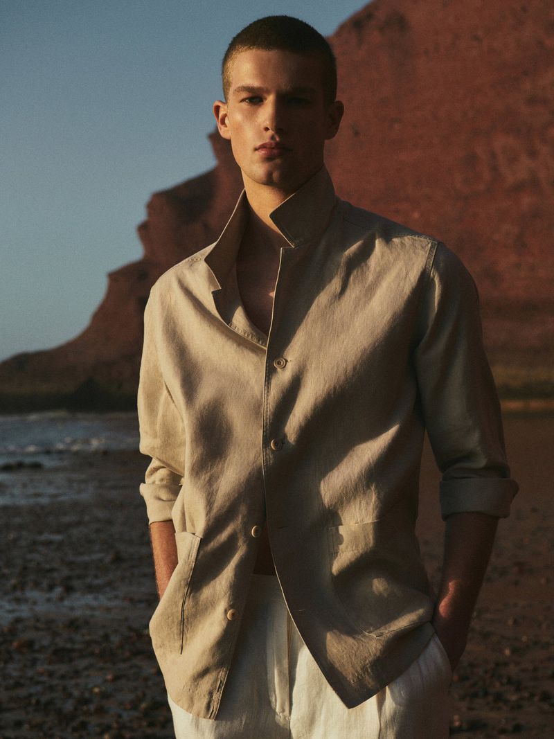 A young adult model poses in an unbuttoned beige linen shirt and white pants against a coastal landscape at golden hour.