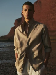 A male model posing on a sandy beach wearing a beige linen shirt and white trousers against rocky cliffs during sunset