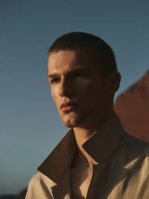 Close-up portrait of a young male model wearing a beige collared shirt against a clear blue sky and rocky landscape.