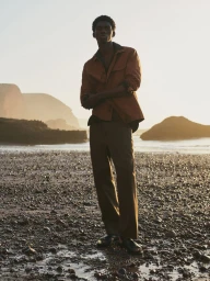 Model Ottawa Kwami standing on a pebble-strewn beach at sunset wearing a rust-colored overshirt and dark trousers