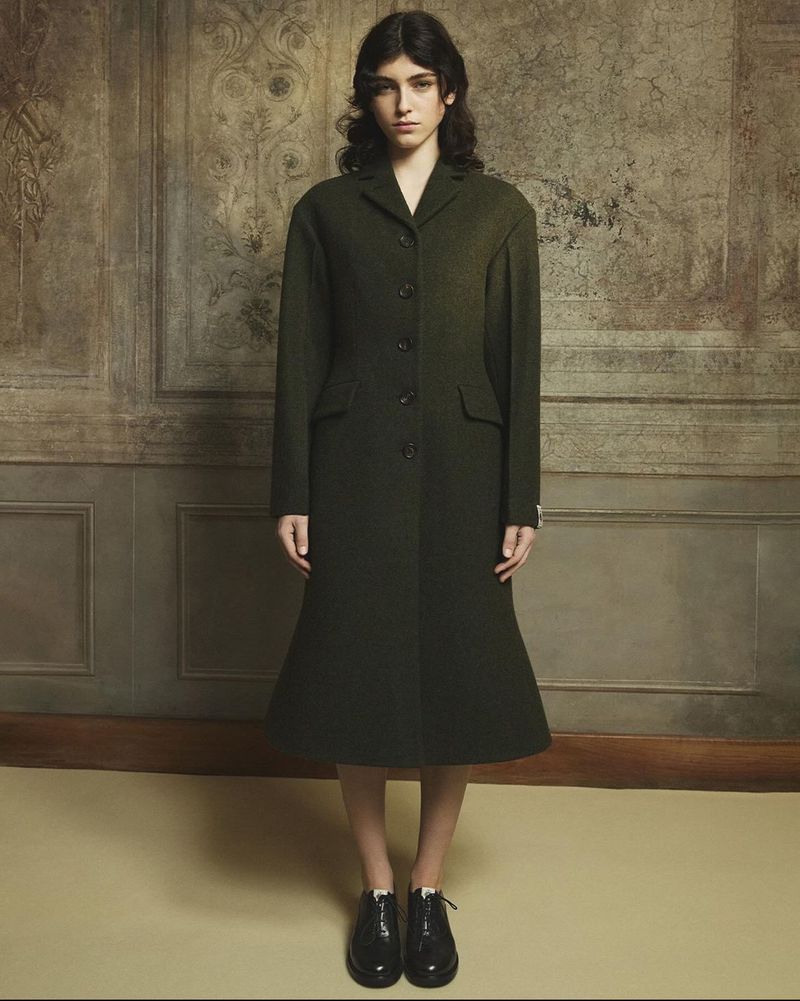 A brunette woman stands in an olive green coat in front of a decorative beige wall, complemented by her dark hair and black shoes.
