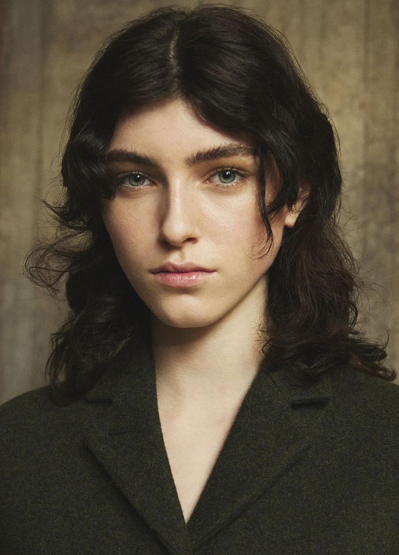 Close-up portrait of a young adult woman with dark wavy hair and green eyes, wearing a dark green coat against a neutral backdrop.