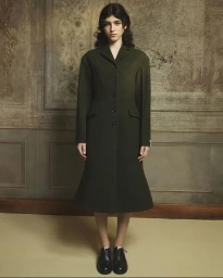 Model standing facing forward wearing a long dark green coat and black shoes against a decorative wall