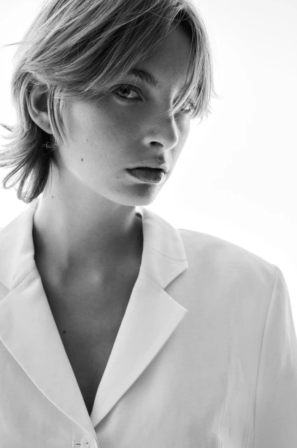 Black and white portrait of a young woman with short hair wearing a white blazer against a bright, neutral background, looking at the camera.