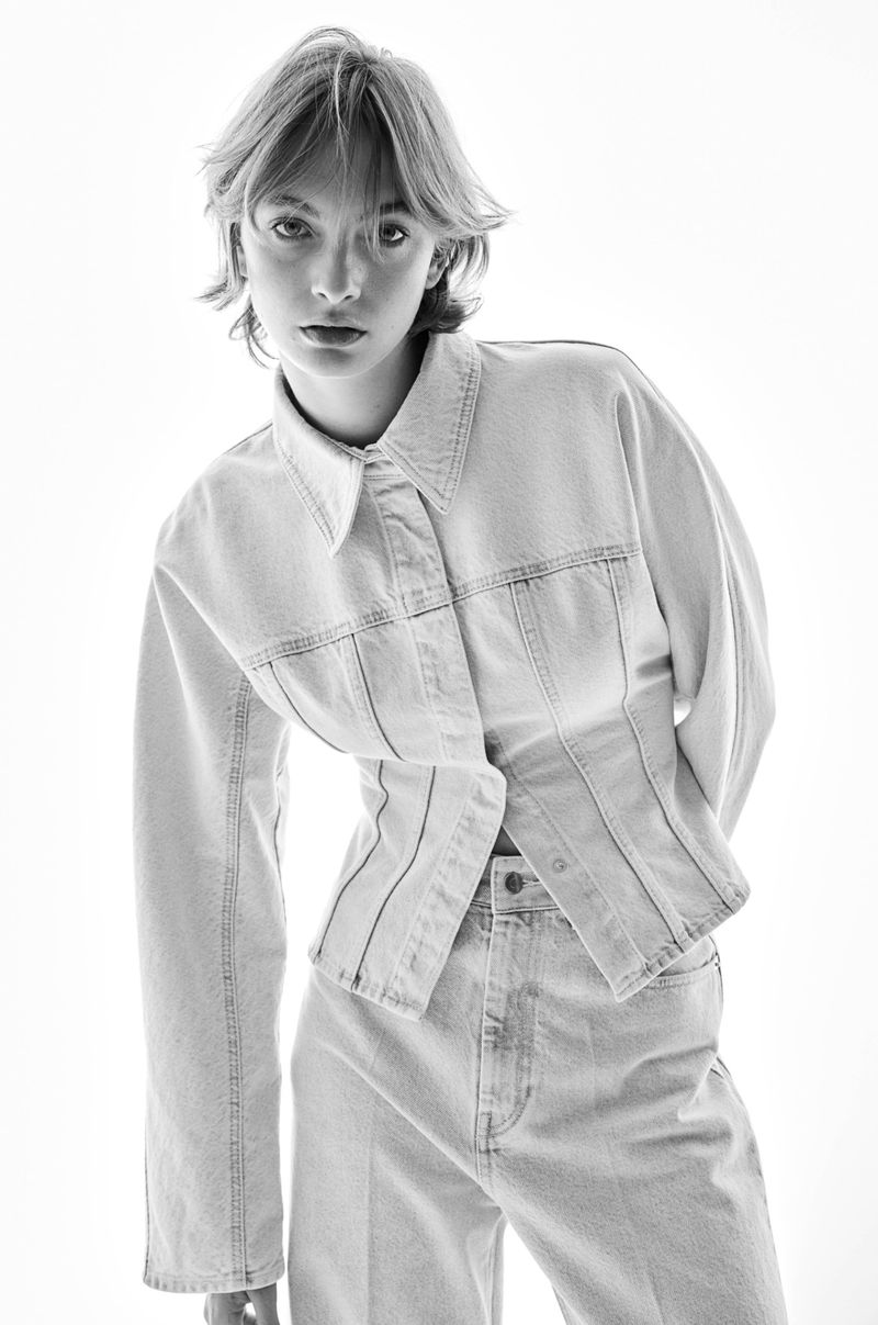 Studio shot of a young adult female model with short blonde hair wearing a denim jacket and light denim jeans against a white backdrop.