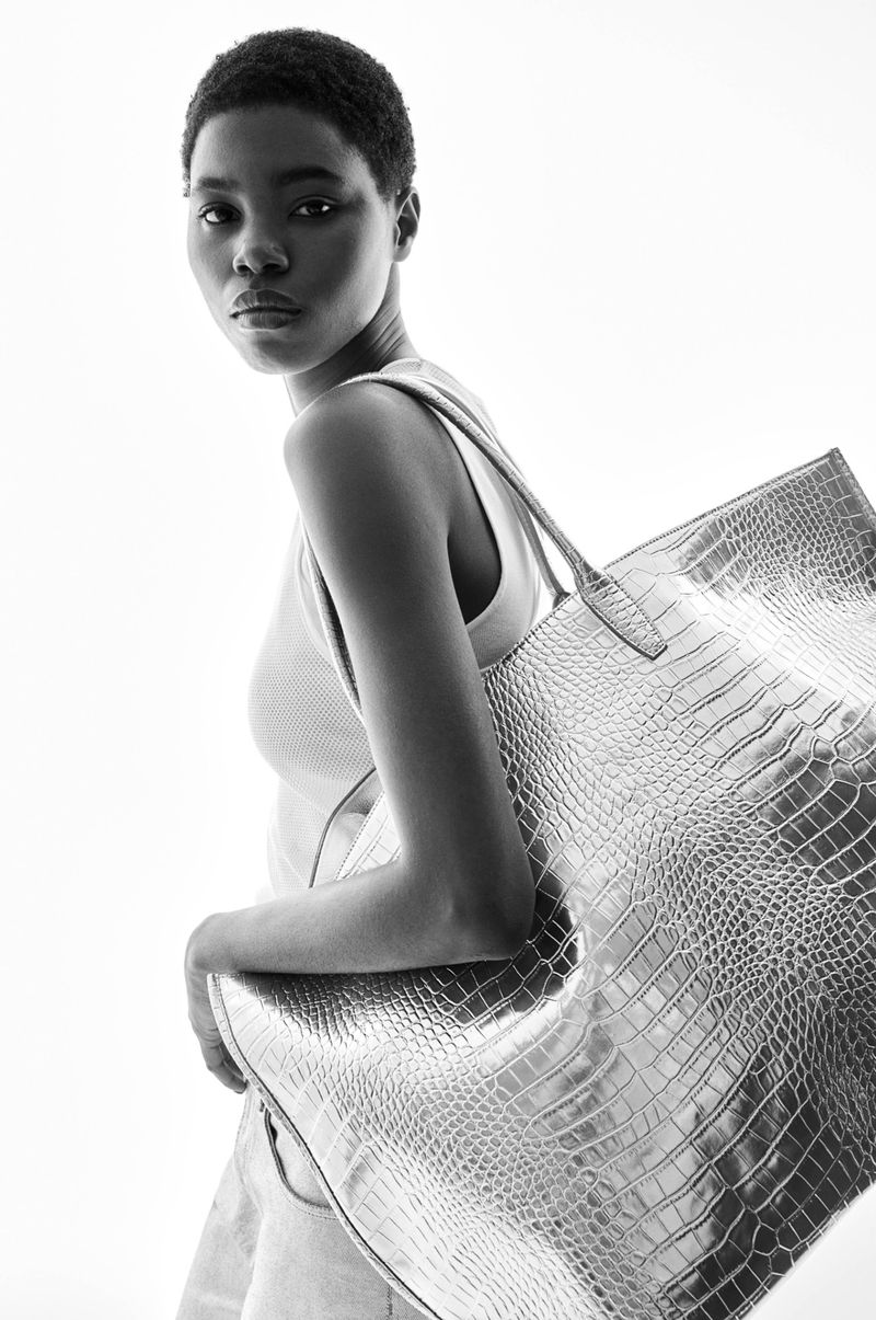 A fashionable Black model showcases a large silver tote bag with a crocodile texture against a bright white background in a studio setting.