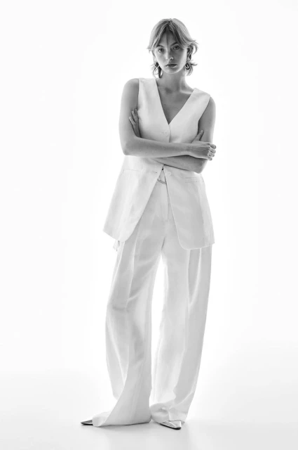 Black and white studio shot of a young adult woman posing in a white sleeveless top and matching wide-leg pants with arms crossed.