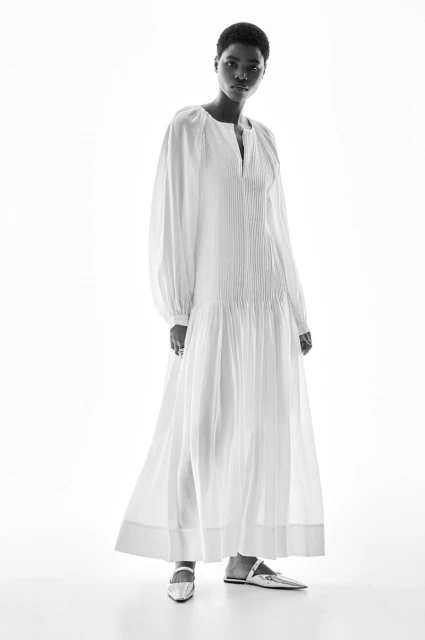 Black woman in a white long-sleeved maxi dress and silver flats posing against a white backdrop in a fashion studio setting.