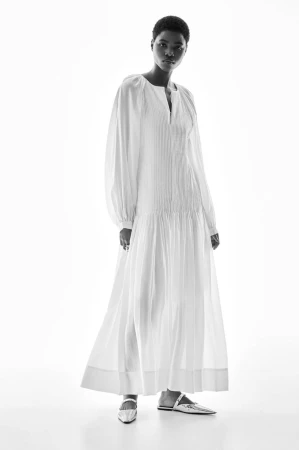 Black woman in a white long-sleeved maxi dress and silver flats posing against a white backdrop in a fashion studio setting.