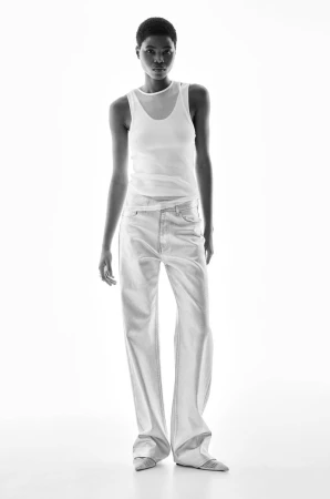 Model in white tank top and light trousers standing against a seamless white background