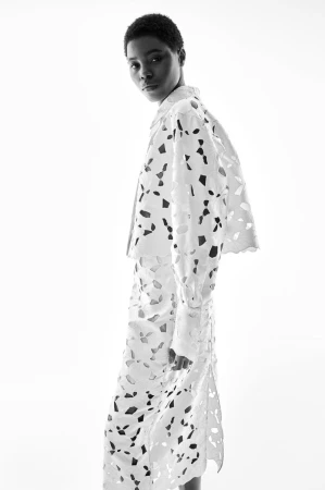 Monochrome image of an African woman in a white laser-cut jacket and skirt, posing in a brightly lit, minimalist studio.