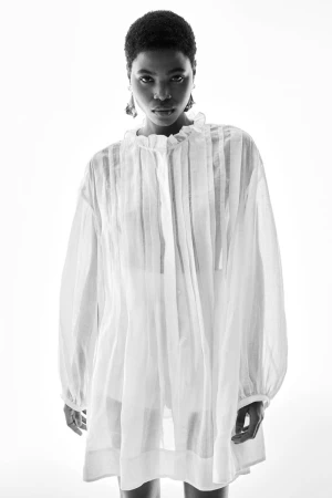 Black woman wearing a sheer white ruffled tunic shirt against a plain white studio background.