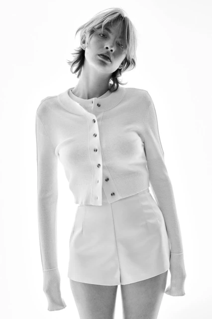 Black and white portrait of a young woman in a white short set looking at the camera with a neutral expression in a bright, studio setting.
