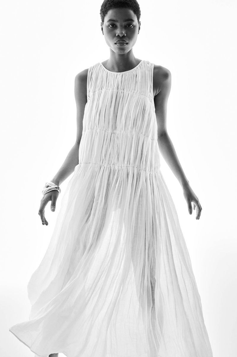 A young, slender adult woman poses in a floor-length white dress and bangle in a studio setting against a bright white background.