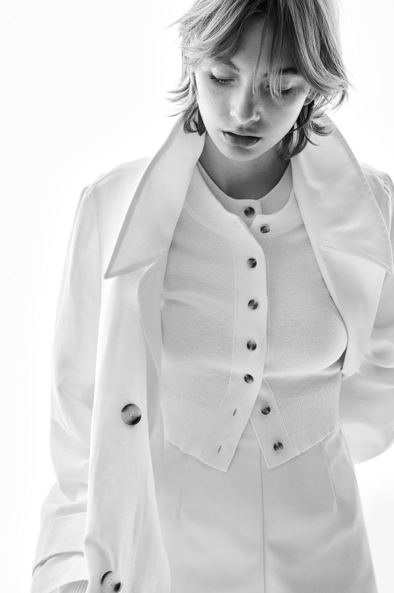 Monochrome portrait of a woman in a white button-up shirt and trench coat looking down in a brightly lit studio setting.