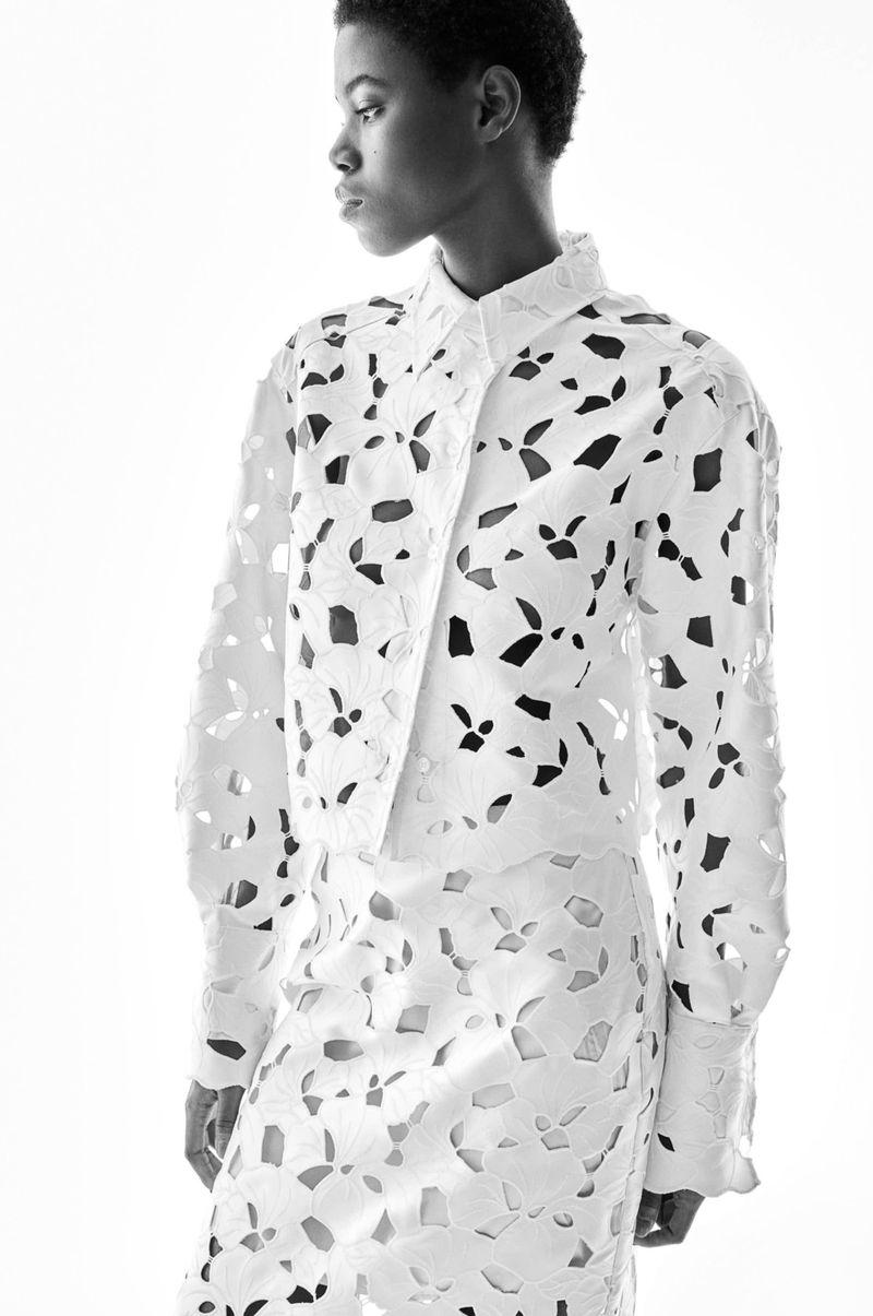Monochrome fashion shot of a Black woman in a white laser-cut shirt and skirt, set against a bright white backdrop in a studio setting.