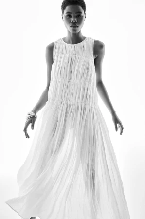 Model wearing a pleated white sleeveless dress against a bright white background for the H&M S/S 2024 campaign.