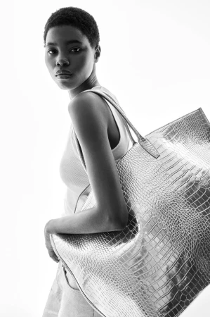 Model with short hair wearing a white tank top carrying a large metallic silver tote bag against a bright white background