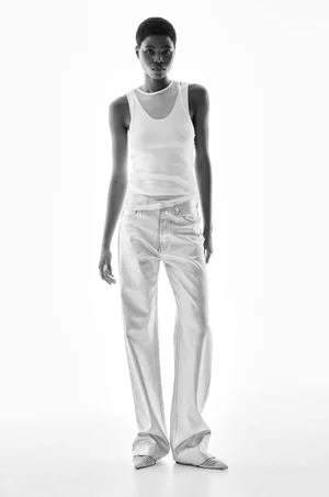 Model in white tank top and light trousers standing against a seamless white background