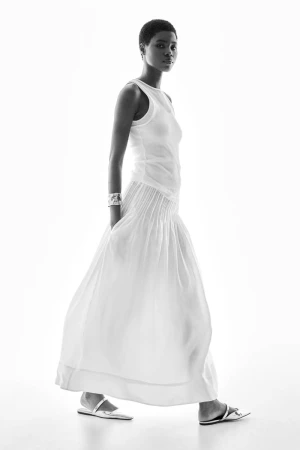 Model wearing a white tank top and long white skirt standing against a bright white background