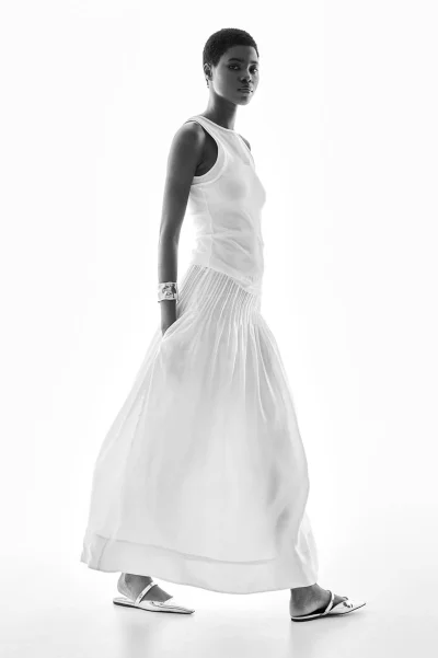 Model wearing a white tank top and long white skirt standing against a bright white background