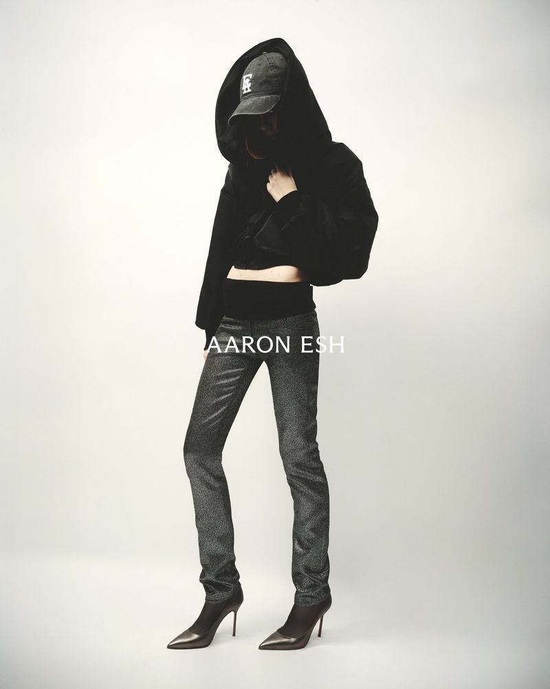 Full-length shot of a woman in a hooded black jacket, grey glittering pants, and heels posing against a white background.