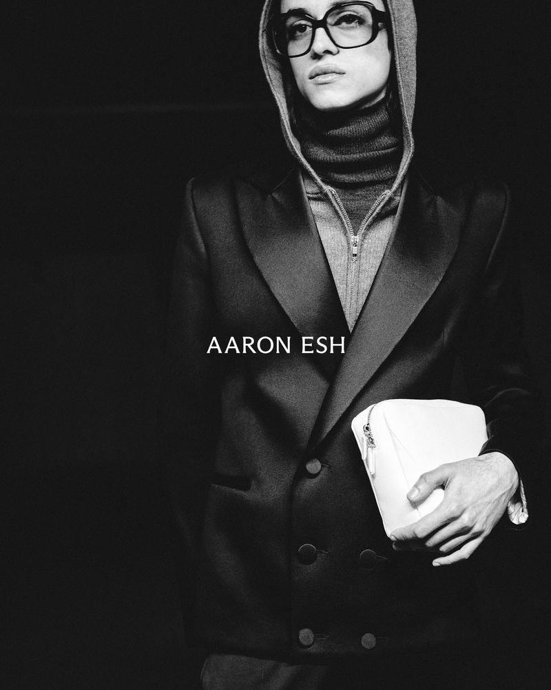 Monochrome studio portrait of a person in glasses and hoodie, wearing a jacket, and holding a white clutch against a dark background.