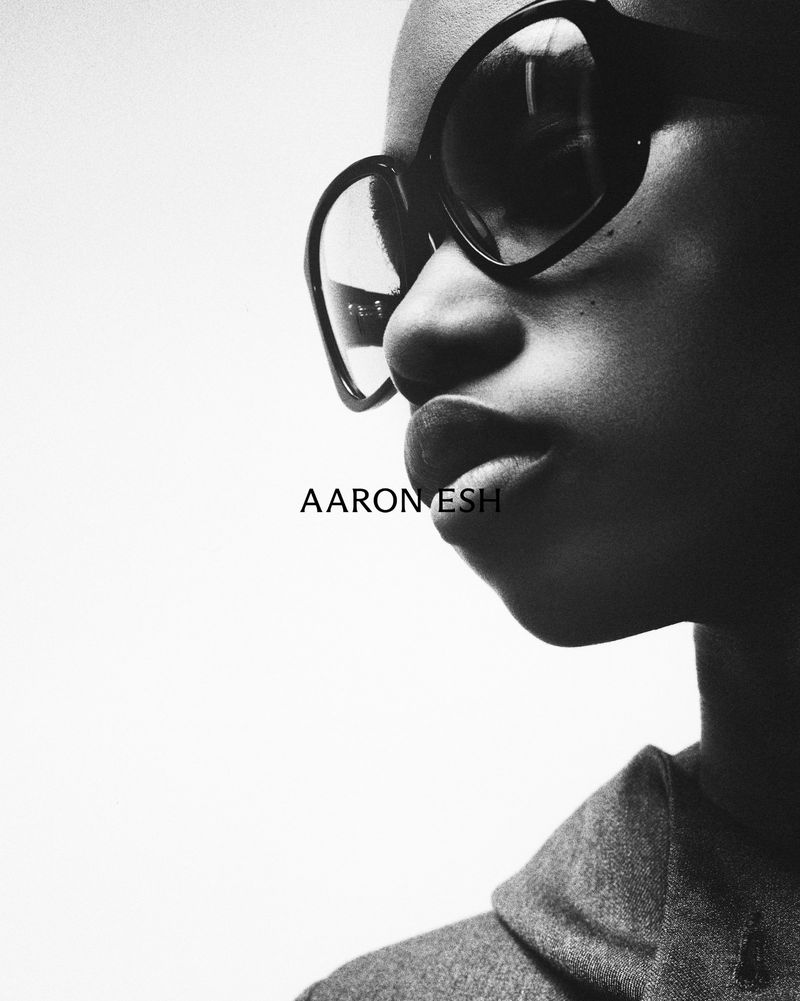 Monochrome portrait of a person with dark skin wearing dark glasses and a grey jacket against a white background in a studio setting.