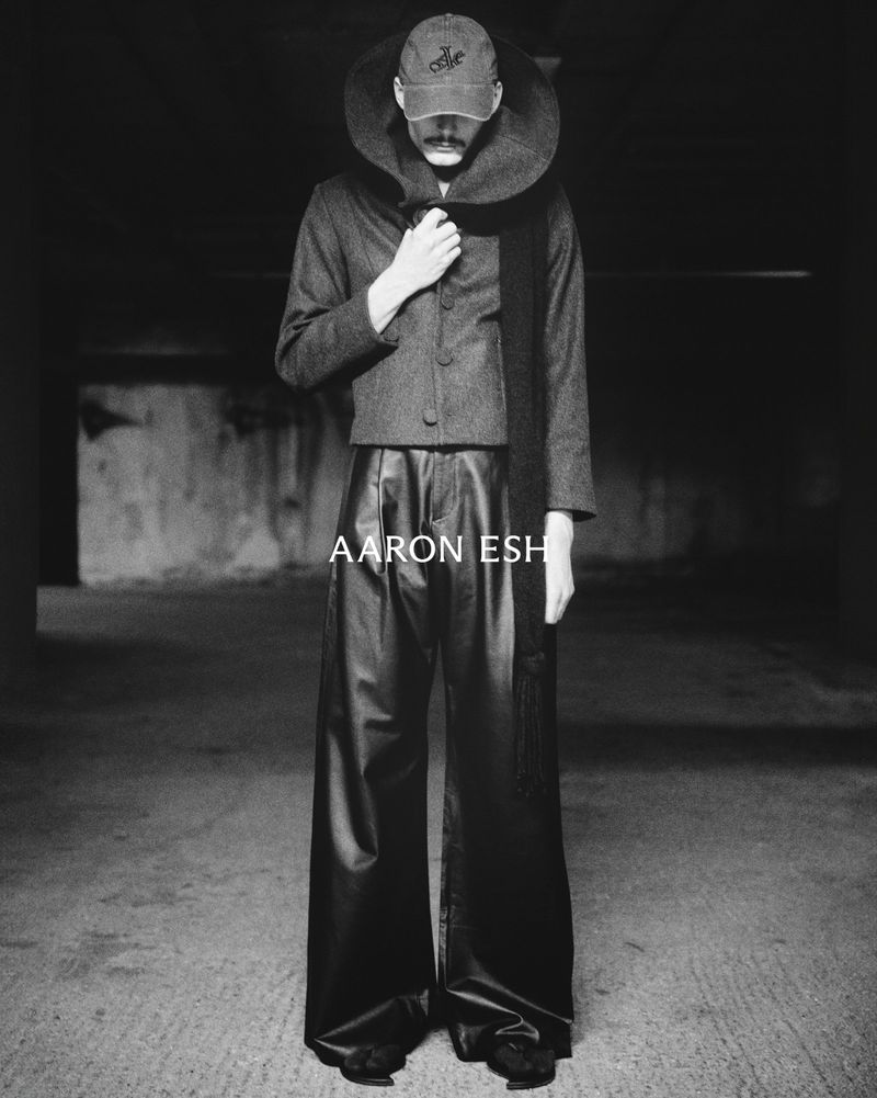 Monochrome portrait of a person wearing oversized leather pants, a wool jacket, and a baseball cap in a dark urban setting.