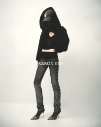 Full-length portrait of a model wearing a black hooded jacket, cap, and patterned trousers against a neutral backdrop.
