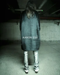 Model standing facing away in a dark textured coat and metallic silver trousers inside a concrete garage.