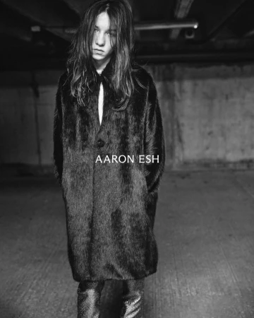 Model in a black textured coat standing in a dim industrial space for Aaron Esh A/W 24