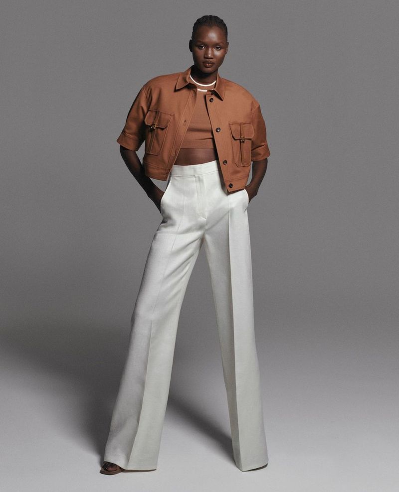 A fashion model poses in a studio wearing a cropped jacket, matching top, and white pants against a grey background, showcasing a modern fashion style.