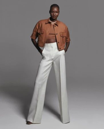 A fashion model poses in a studio wearing a cropped jacket, matching top, and white pants against a grey background, showcasing a modern fashion style.