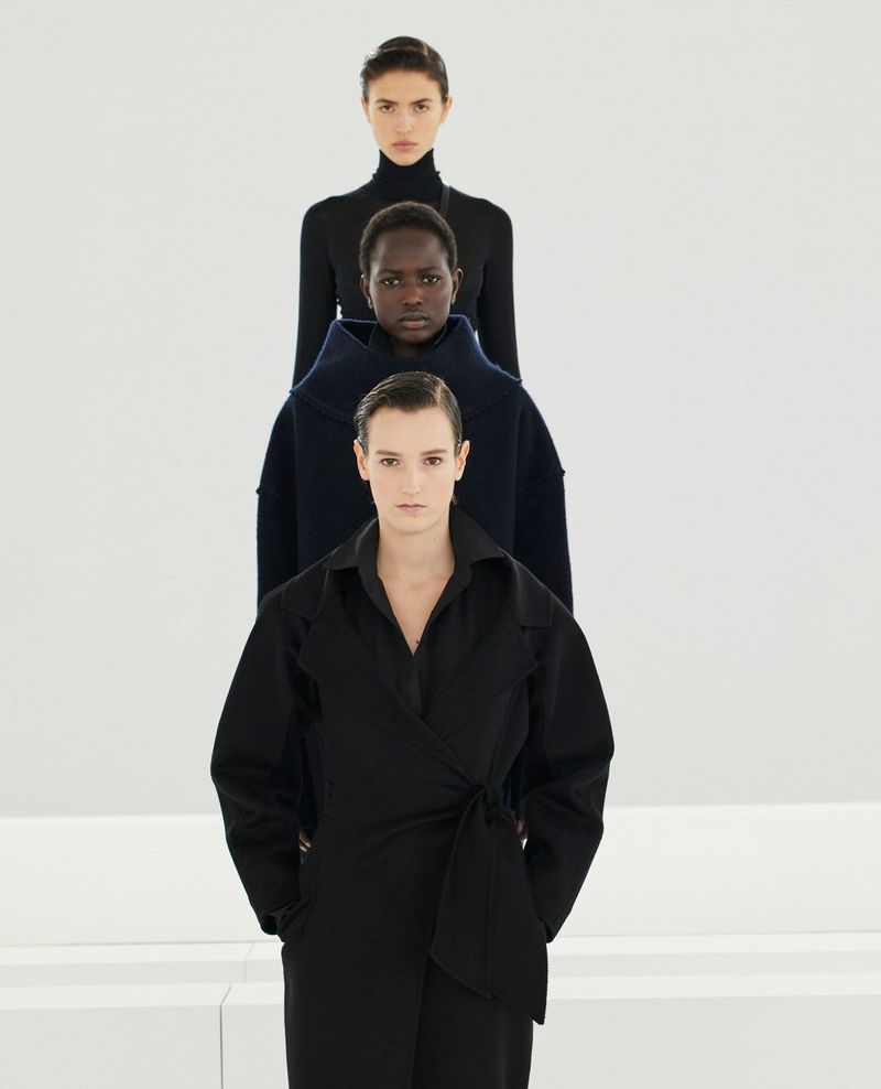 Three fashion models stand in a line against a white background, each wearing a different style of dark-colored, minimalist clothing.