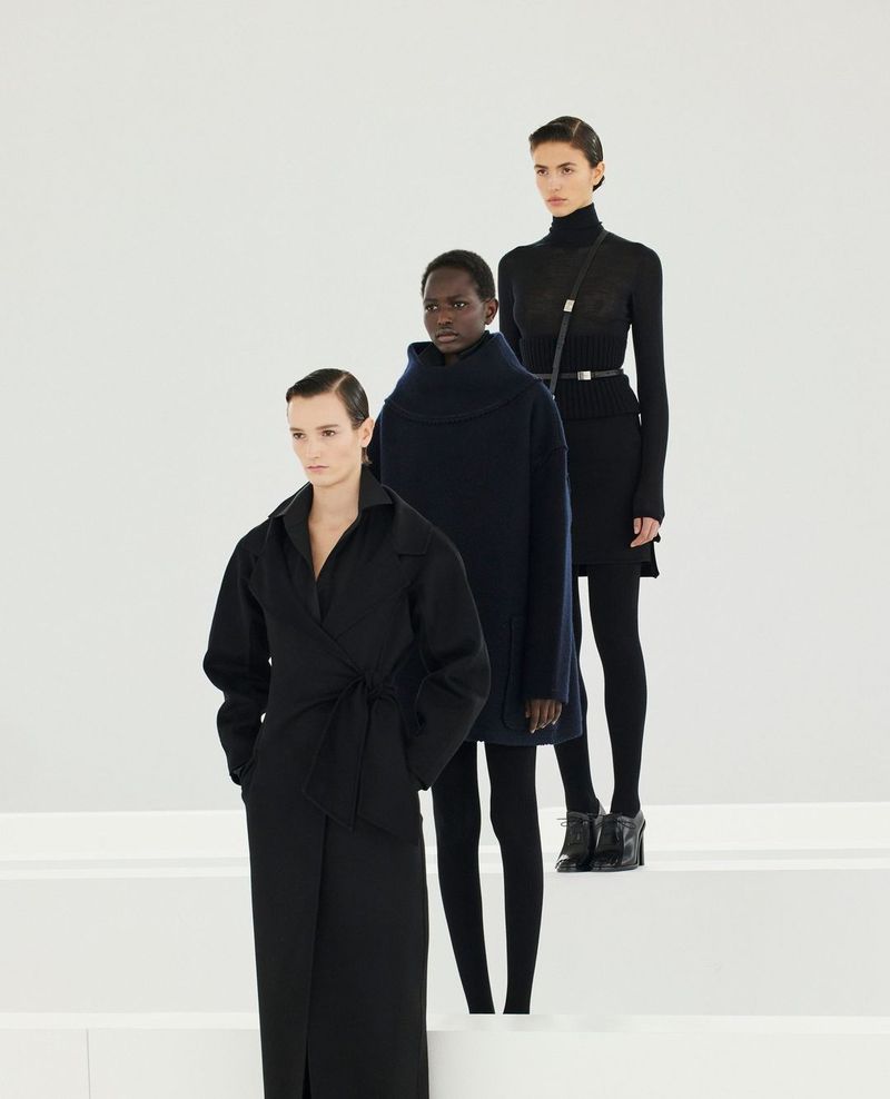 Three female fashion models in black clothing pose in a bright studio setting on white steps, presenting a modern, minimalist aesthetic.