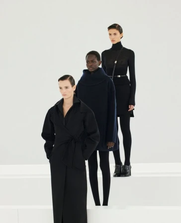Three female fashion models in black clothing pose in a bright studio setting on white steps, presenting a modern, minimalist aesthetic.