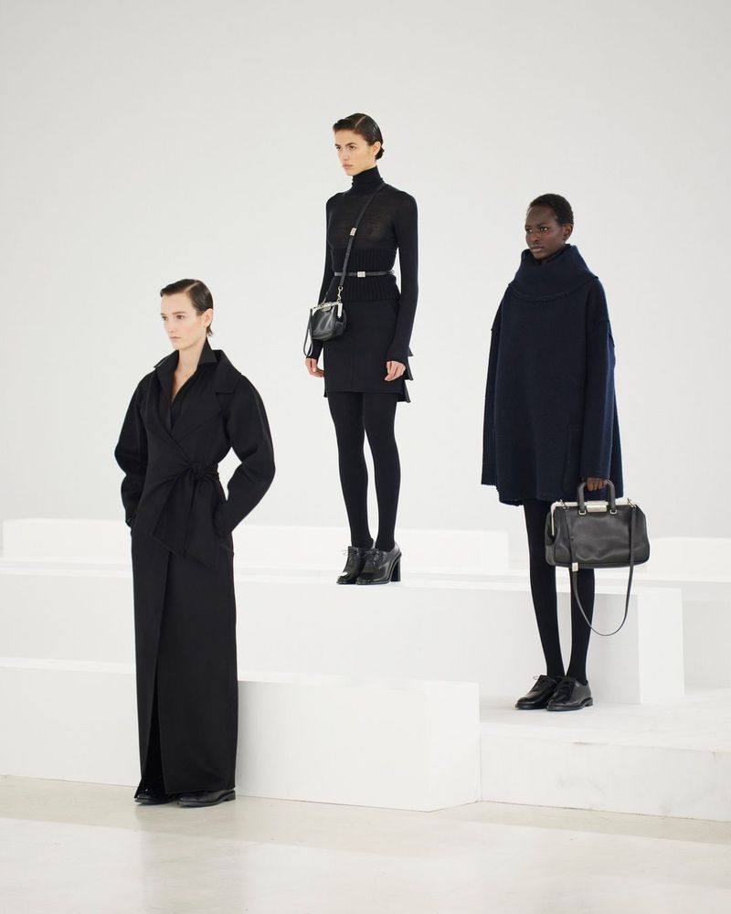 Three fashion models showcase black outfits and purses on a white staircase in a studio setting, highlighting minimalist and modern styles.